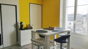 A small kitchen with a white table, two bar stools, and a window with a view of buildings. - Vittel Loc's : Le 214 Classé 3 Osez L'expérience! (Vittel)