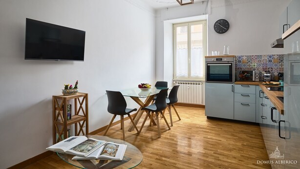 A modern kitchen with a flat-screen TV, a dining area with a glass table, and a view of the outside through a window. - Domus Alberico 11 (Vaticano) (Roma)