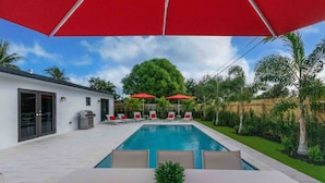 Un espace piscine extérieur moderne, avec une table et des chaises blanches, des parasols rouges et une pelouse bien entretenue.