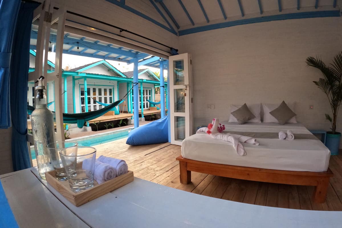 A bedroom with a bed, a wooden table with a glass of water and a bottle, a hammock outside, and a view of a pool and blue buildings.