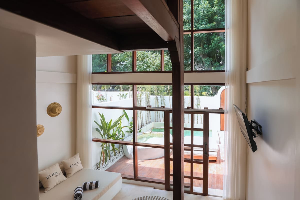 A room with a large window, a sofa with cushions, and a view of an outdoor pool area.