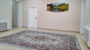 A spacious room with a large patterned rug, a white door, and a wall-mounted picture of a landscape. - Saroy Palace Hotel (Qarshi)