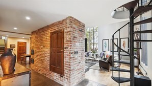 Een ruime woonkamer met een accentmuur van baksteen, een trap en een groot raam dat voor natuurlijk licht zorgt.