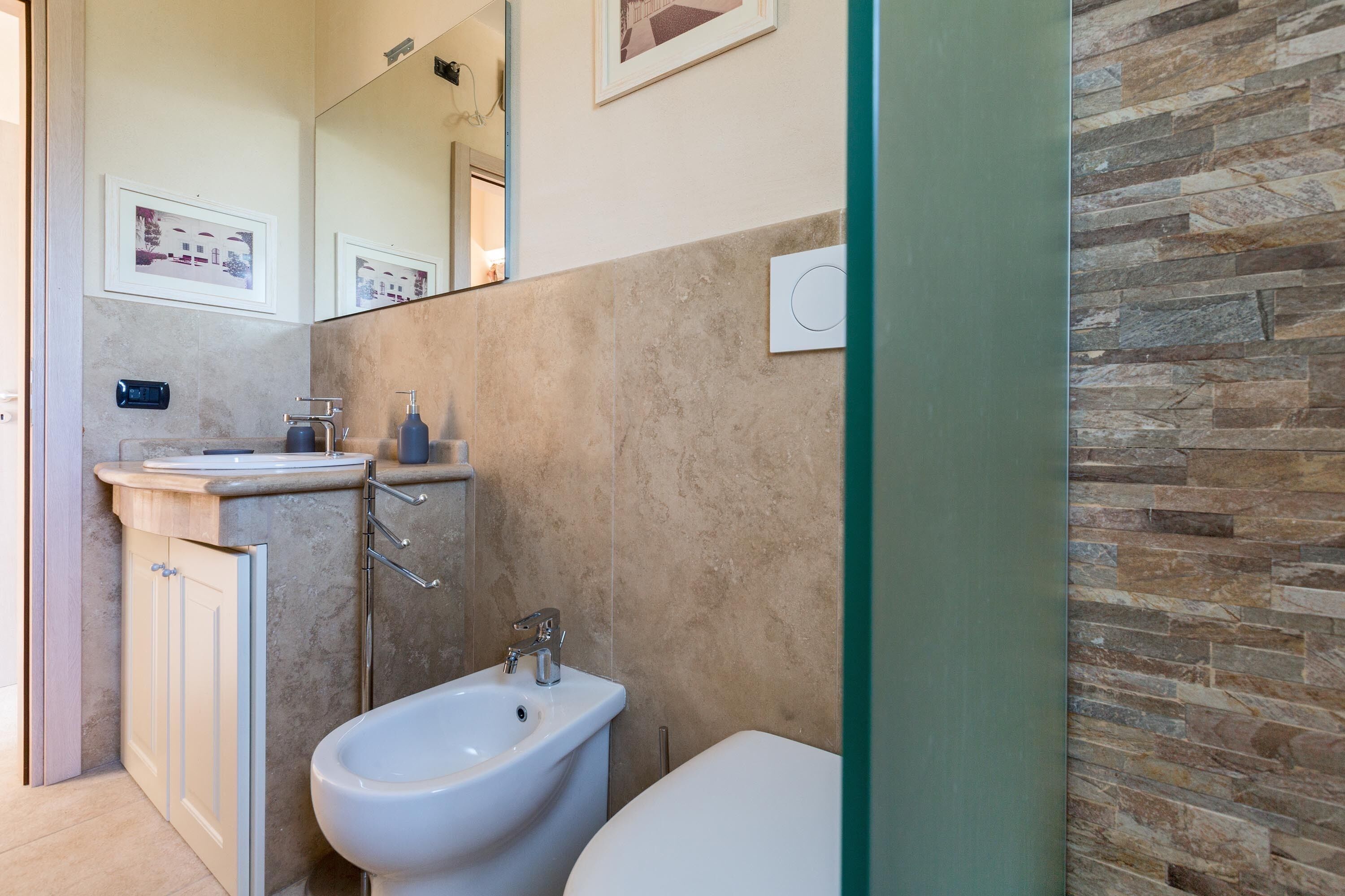 Une salle de bain équipée d’un bidet, d’un lavabo et de toilettes. Un miroir se trouve au-dessus du lavabo et un tableau encadré est accroché au mur.