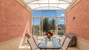 A sunroom with a glass table and chairs, a vase with flowers, and a view of greenery outside. - 3536 Villa Le Majole by Perle di Puglia (Manduria)