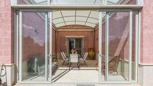 A glass door entrance to a covered patio area with a table and chairs, a large vase, and a wicker chair. - 3536 Villa Le Majole by Perle di Puglia (Manduria)