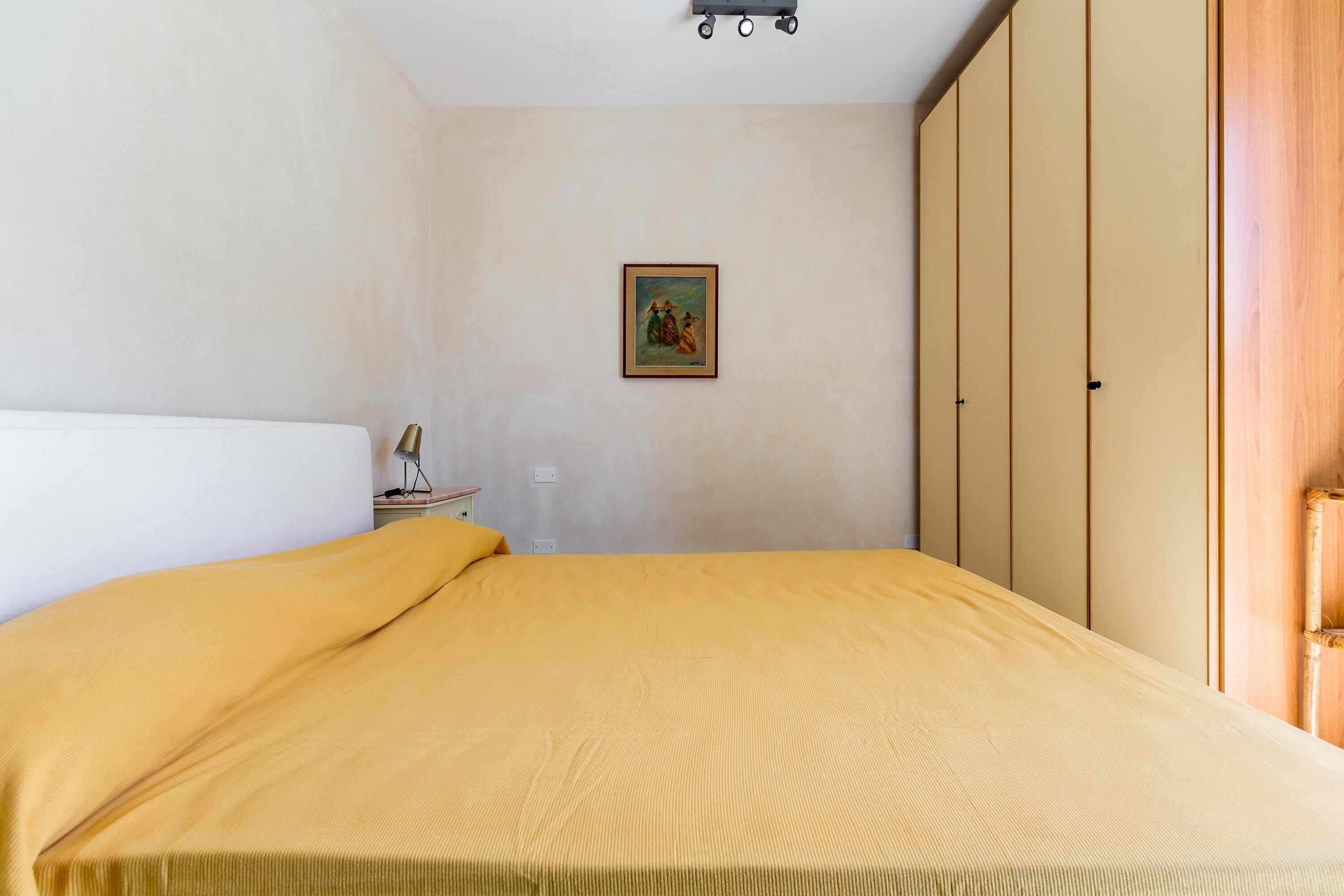 A bedroom with a bed, a nightstand, a lamp, a wardrobe, and a framed picture on the wall.