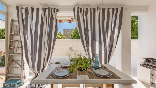 A dining area with a table set for two, striped curtains, a small plant, and a grill in the background. - 3490 Villetta Boschetto by Barbarhouse (Maruggio)