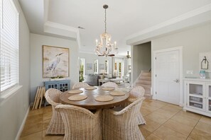 A dining area with a round table, wicker chairs, a chandelier, and a painting of a bird.