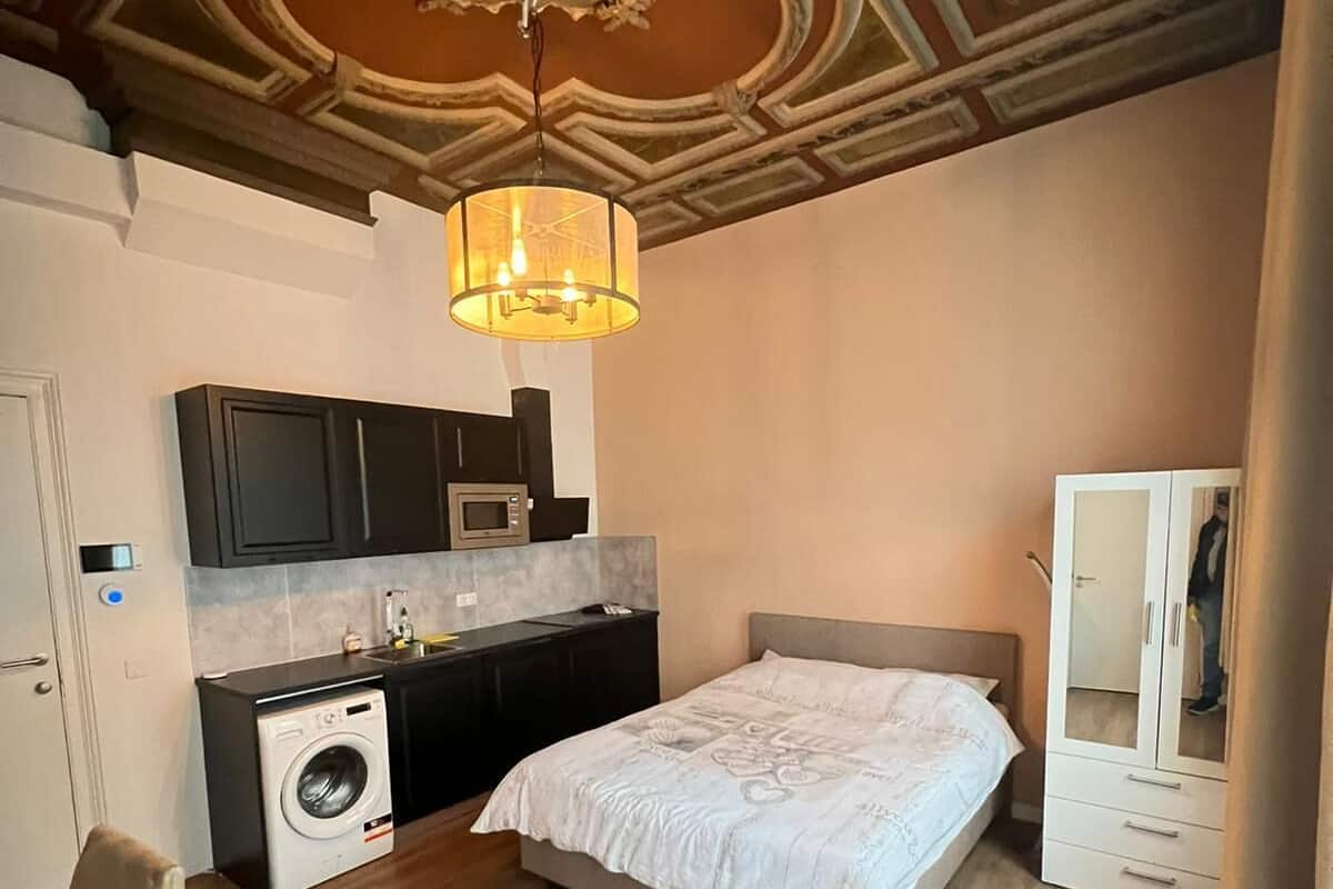 A bedroom with a bed, a washing machine, a kitchenette, and a mirror.