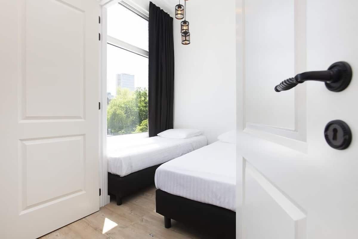A room with two beds, a white door, and a view of greenery outside.