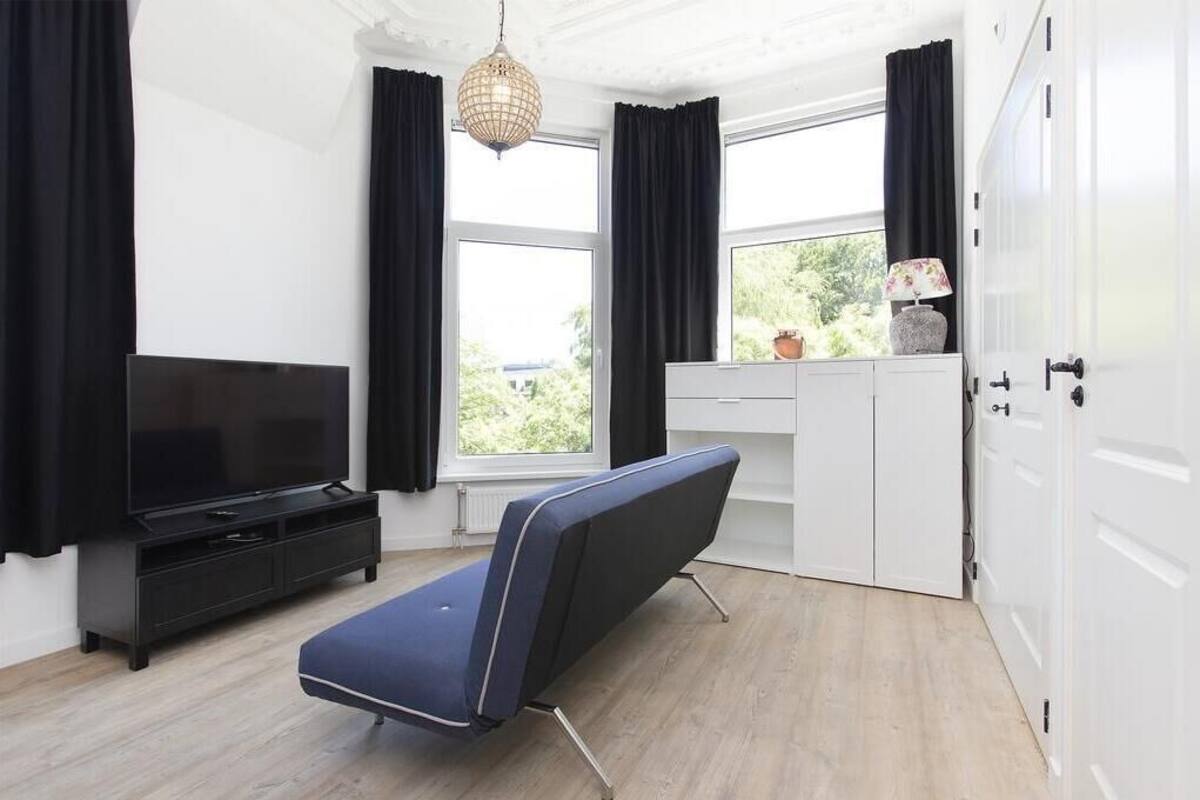 A modern room with a flat-screen TV, a blue bench, a white desk, and a large window with black curtains.