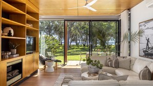 Una sala de estar moderna con un amplio ventanal corredera que da a un jardín, un sofá, un televisor y estanterías de madera.