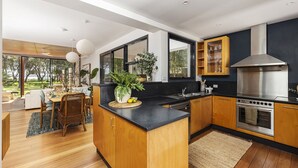 Una cocina moderna con armarios de madera, encimera negra y electrodomésticos de acero inoxidable. Hay una zona de comedor con mesa y sillas, y un ventanal que ofrece vistas a la vegetación.
