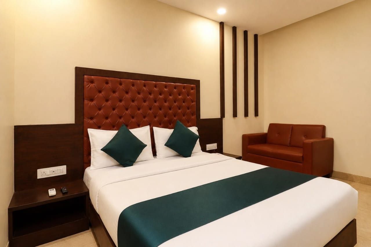A hotel room with a large bed, a brown headboard, a brown sofa, and a nightstand with a phone and charger.