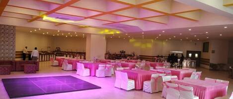Un salón amplio con un escenario, filas de mesas con manteles rosas y sillas blancas.