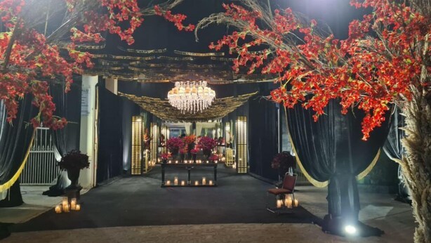 An indoor venue with a central table surrounded by candles, draped with golden fabric, and flanked by red flowering trees.