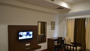 A hotel room with a television mounted on the wall, a desk with a chair, and a window with curtains.