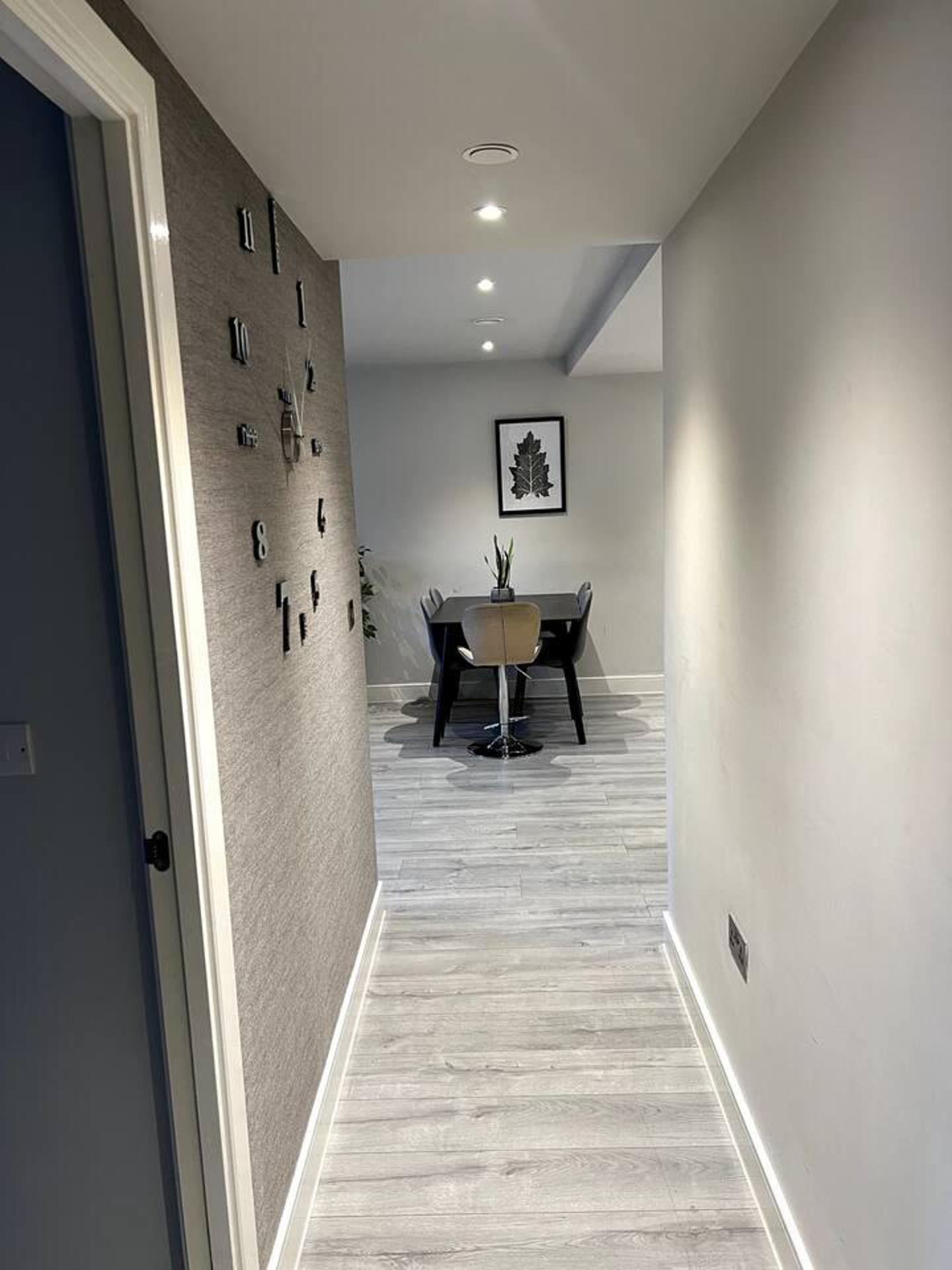 A hallway with a dining area at the end, featuring a table and chairs, a framed picture on the wall, and a clock on the left wall.