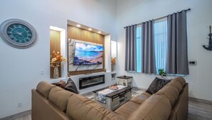 A modern living room with a flat-screen TV, a sofa, a coffee table, and a window with curtains. - Renovated Naples Beach Home w/ Pool & Lanai (Naples)