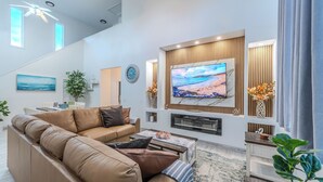 A modern living room with a large flat-screen TV, a brown leather sofa, a wooden accent wall, and a decorative plant. - Renovated Naples Beach Home w/ Pool & Lanai (Naples)