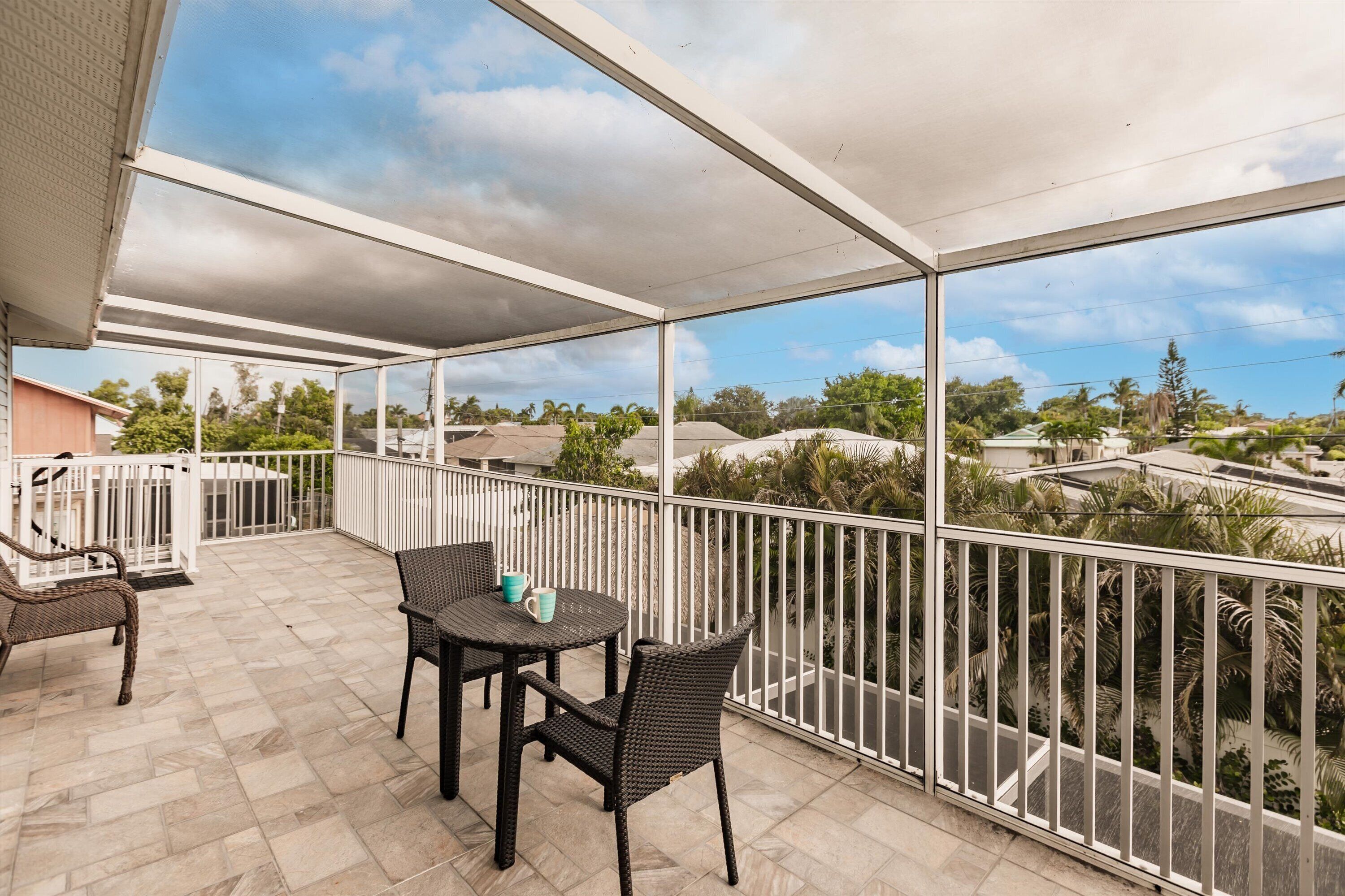 A covered balcony with a table and chairs, offering a view of surrounding houses and palm trees.
