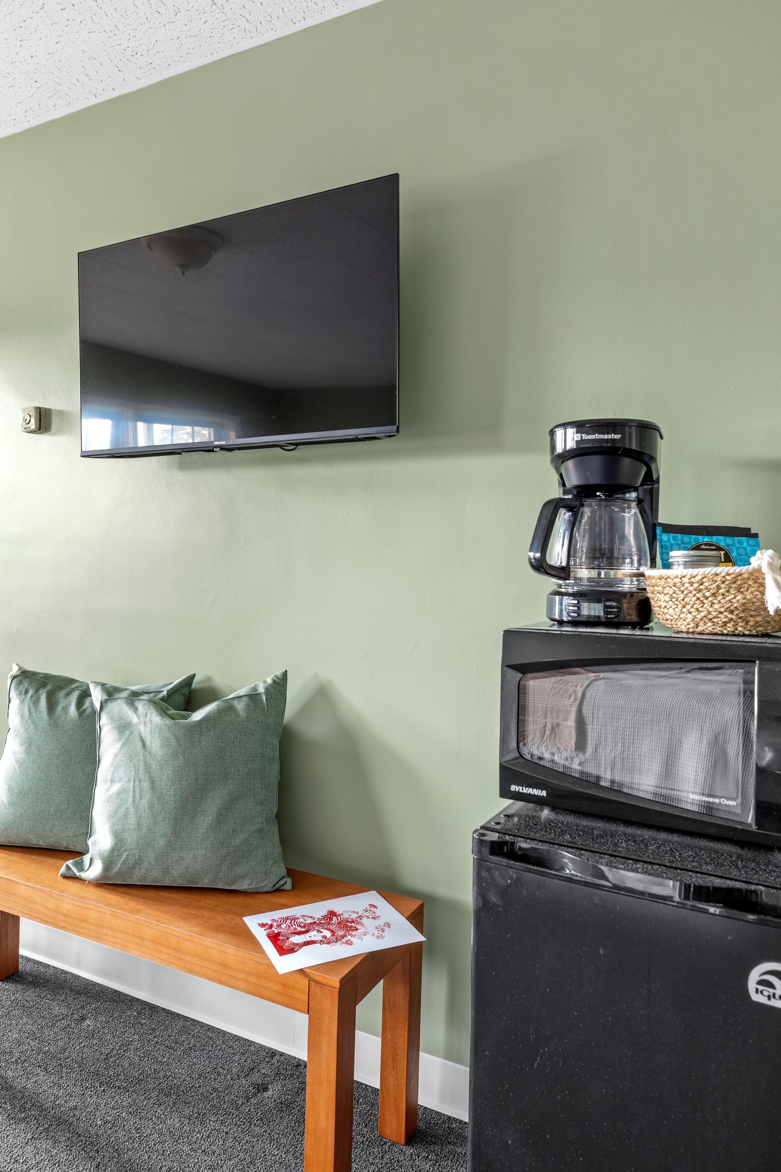 Habitación con televisión de pantalla plana en la pared, cafetera, microondas y un banco con almohadones.
