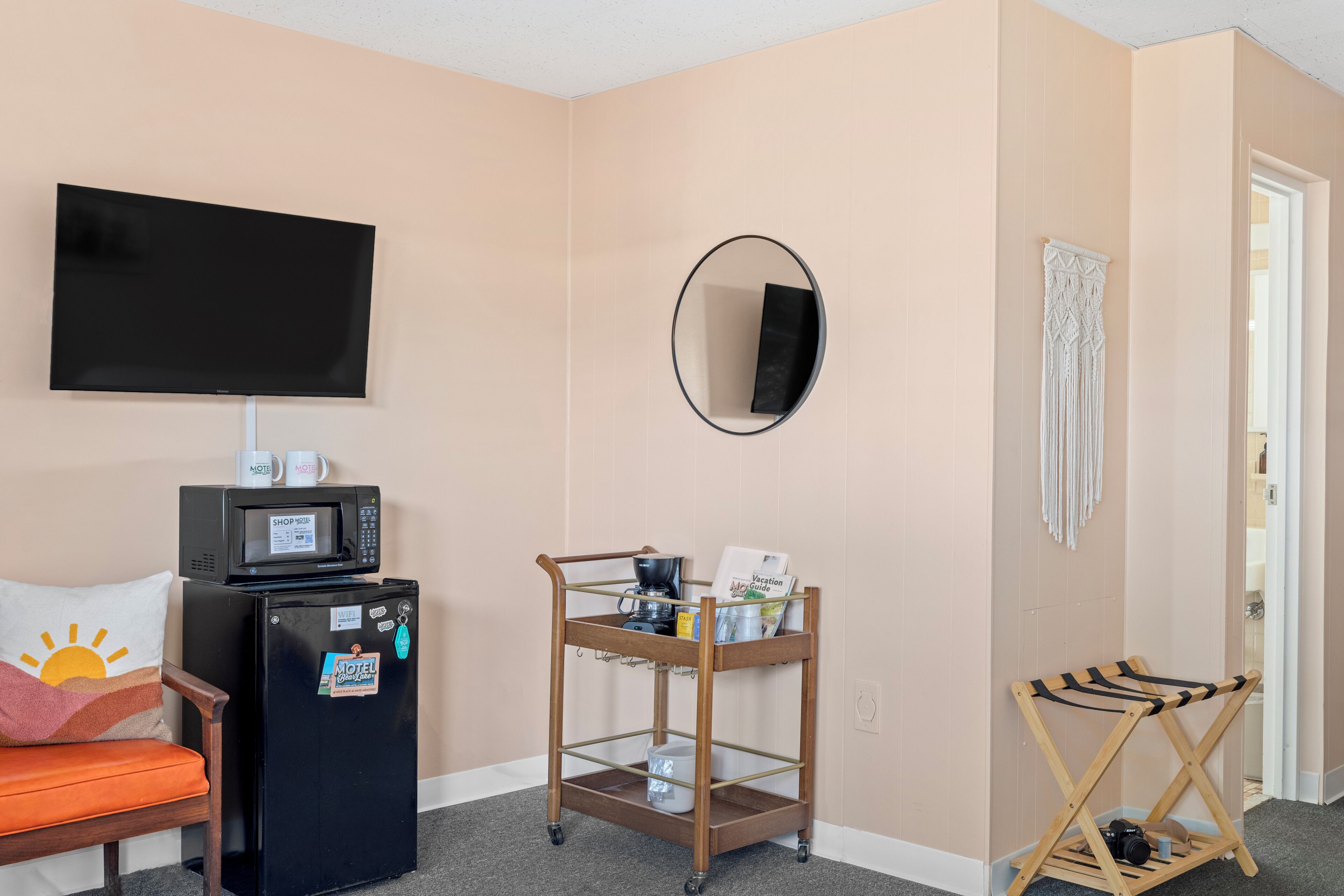 A hotel room with a flat-screen TV, a minibar, a wooden cart with amenities, a mirror, and a door.
