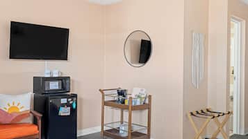 A hotel room with a flat-screen TV, a minibar, a wooden cart with amenities, a mirror, and a door.