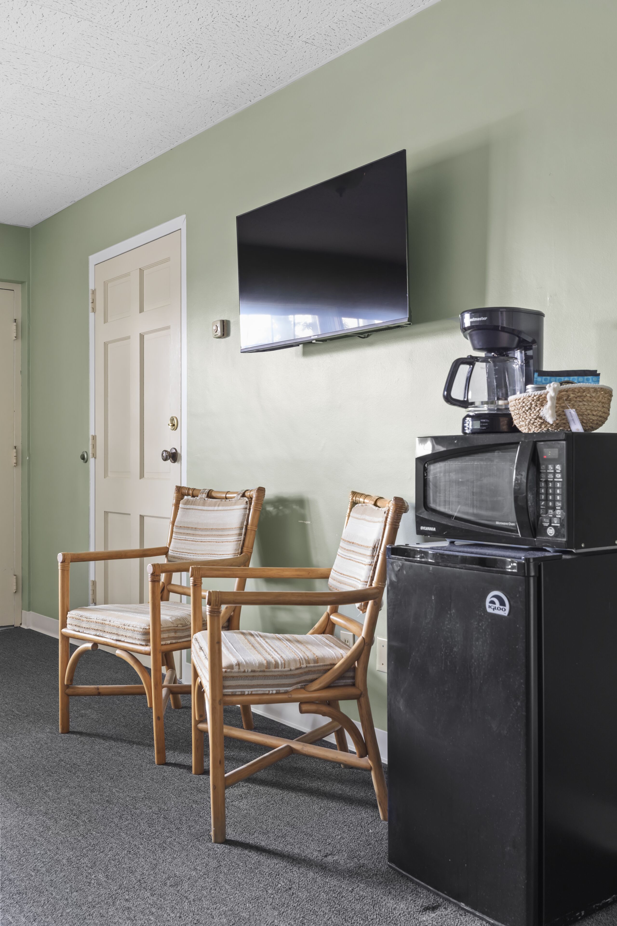 A room with a television mounted on the wall, a microwave, and a coffee maker on a counter. There are two wicker chairs and a door with a white frame.