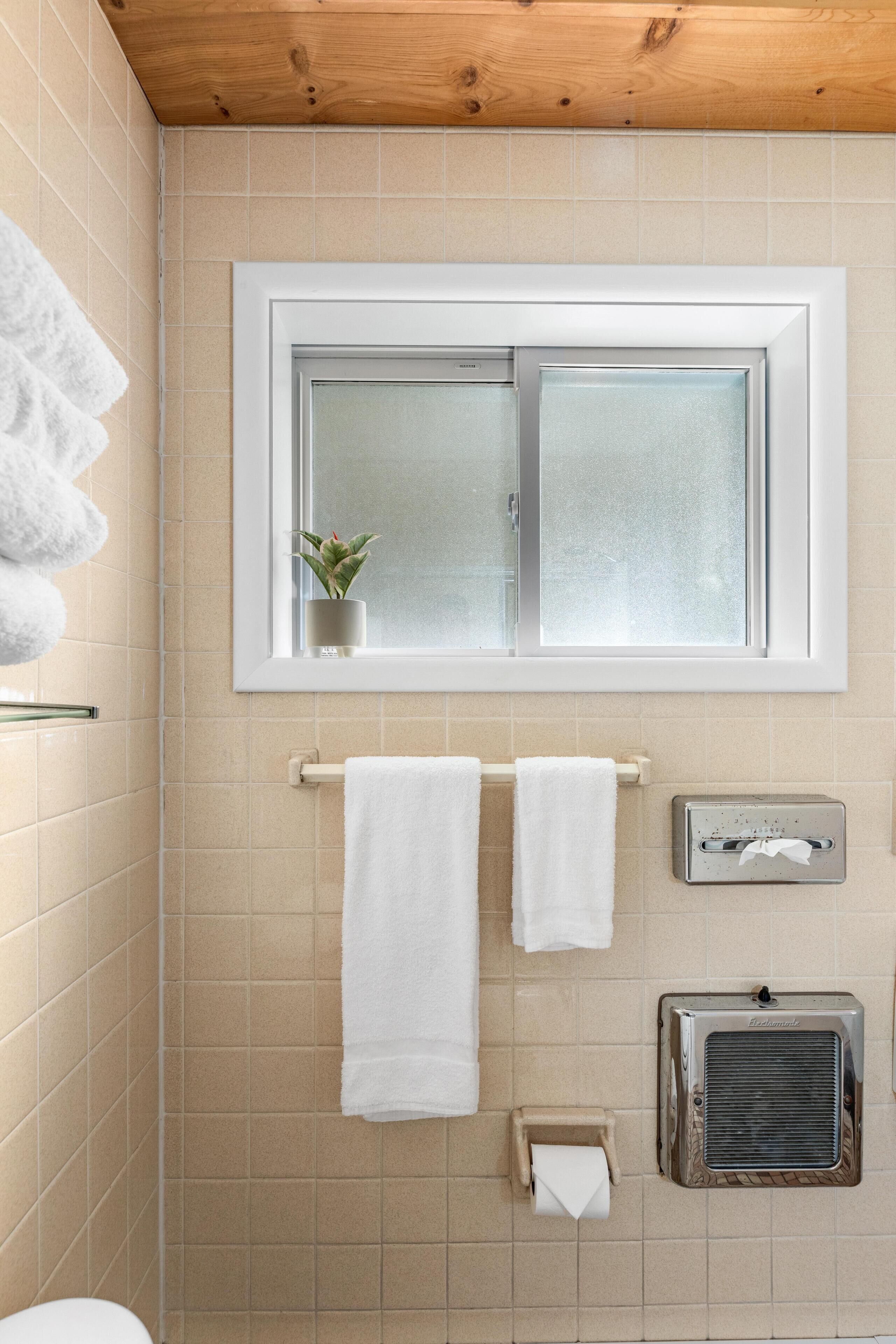 A bathroom with beige tiles, a window with frosted glass, a potted plant, two white towels, a paper towel dispenser, and a wall-mounted heater.