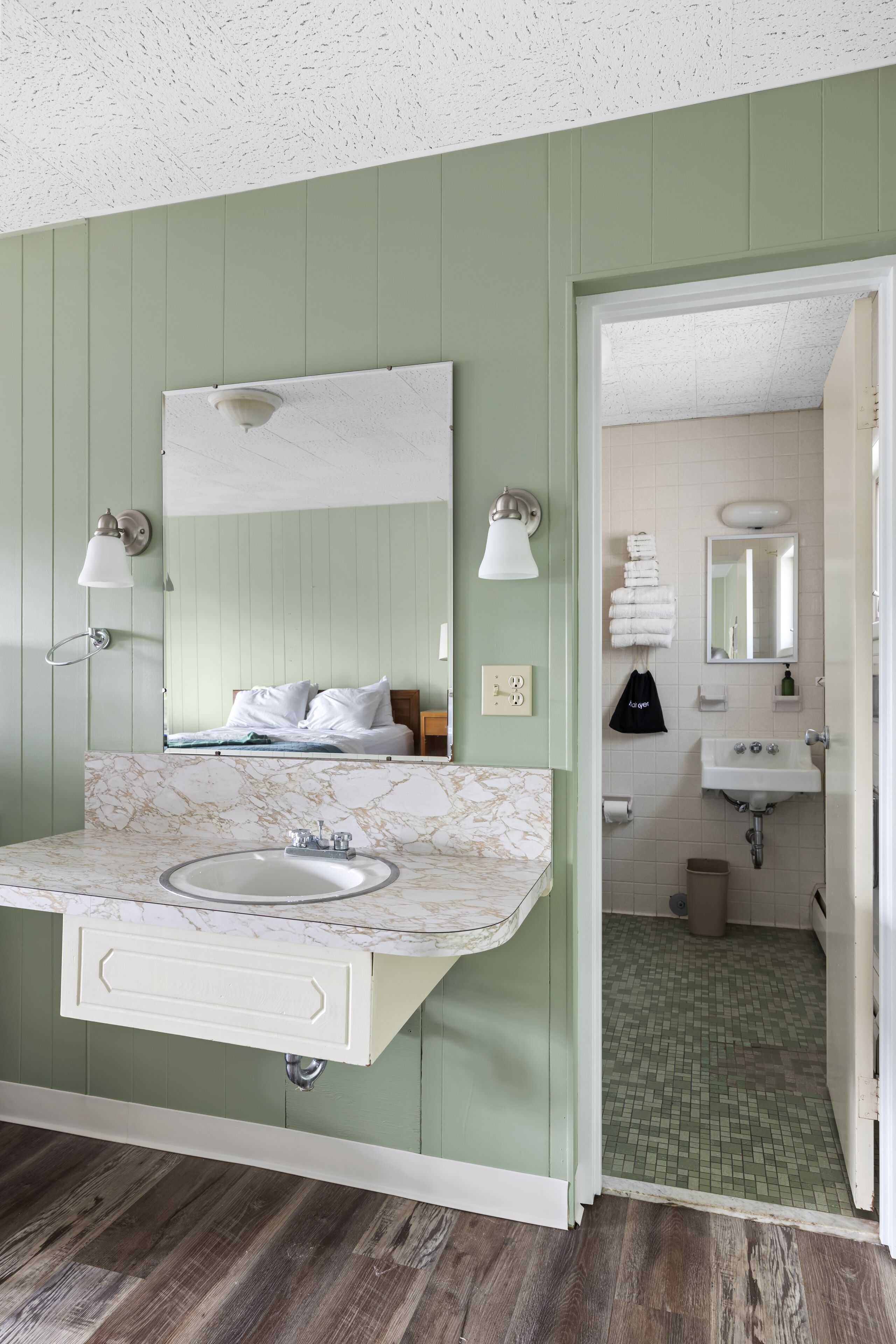A bathroom with a green wall, a marble sink, and a mirror. There is a bed with white bedding in the background.
