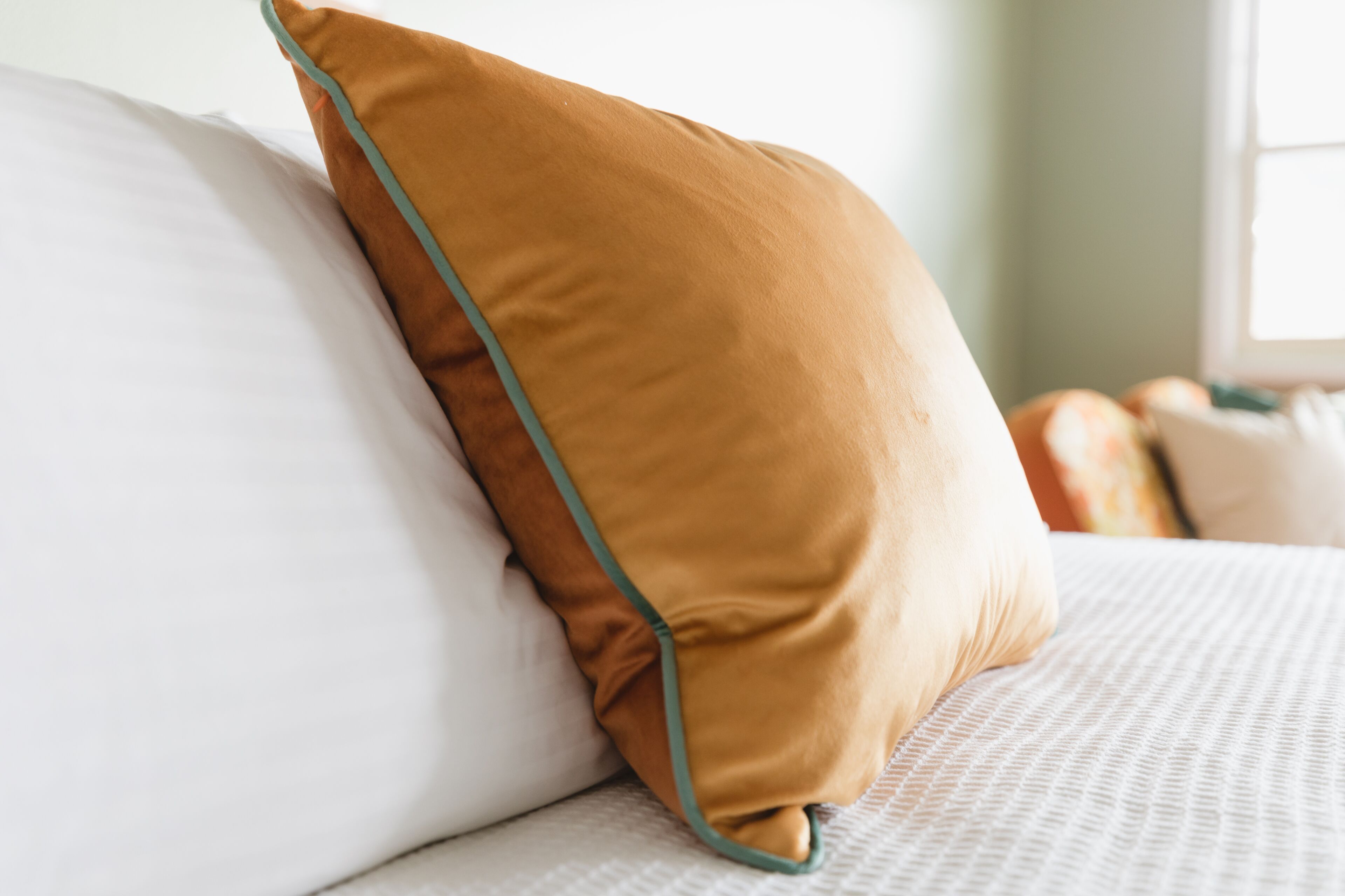 Een bed met witte lakens en een mosterdkleurig kussen met groene biezen.
