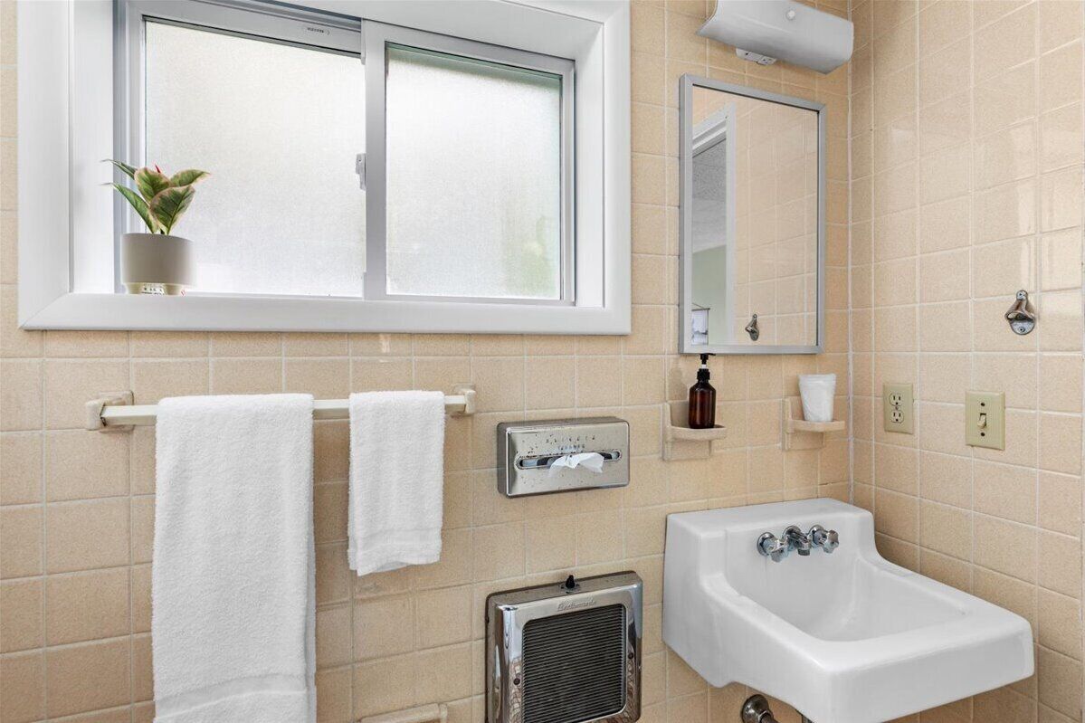 Un baño con ventana, un lavabo, un espejo, un perchero para toallas, un calentador y un estante pequeño con artículos de aseo.