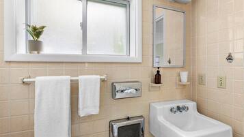 Un baño con ventana, un lavabo, un espejo, un perchero para toallas, un calentador y un estante pequeño con artículos de aseo.