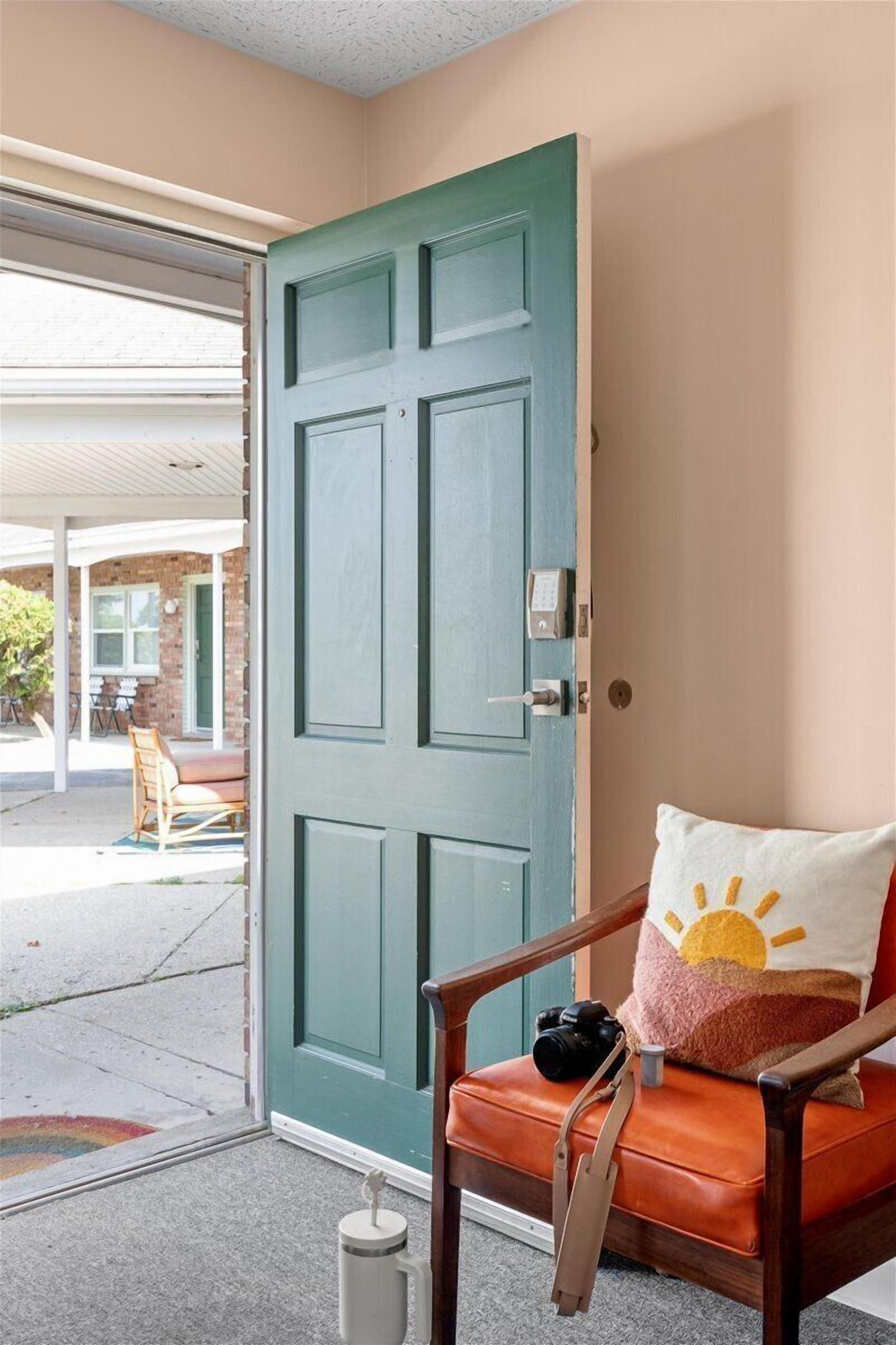 A room with a teal door, a wooden chair with a cushion, and a small table with a camera and a mug.