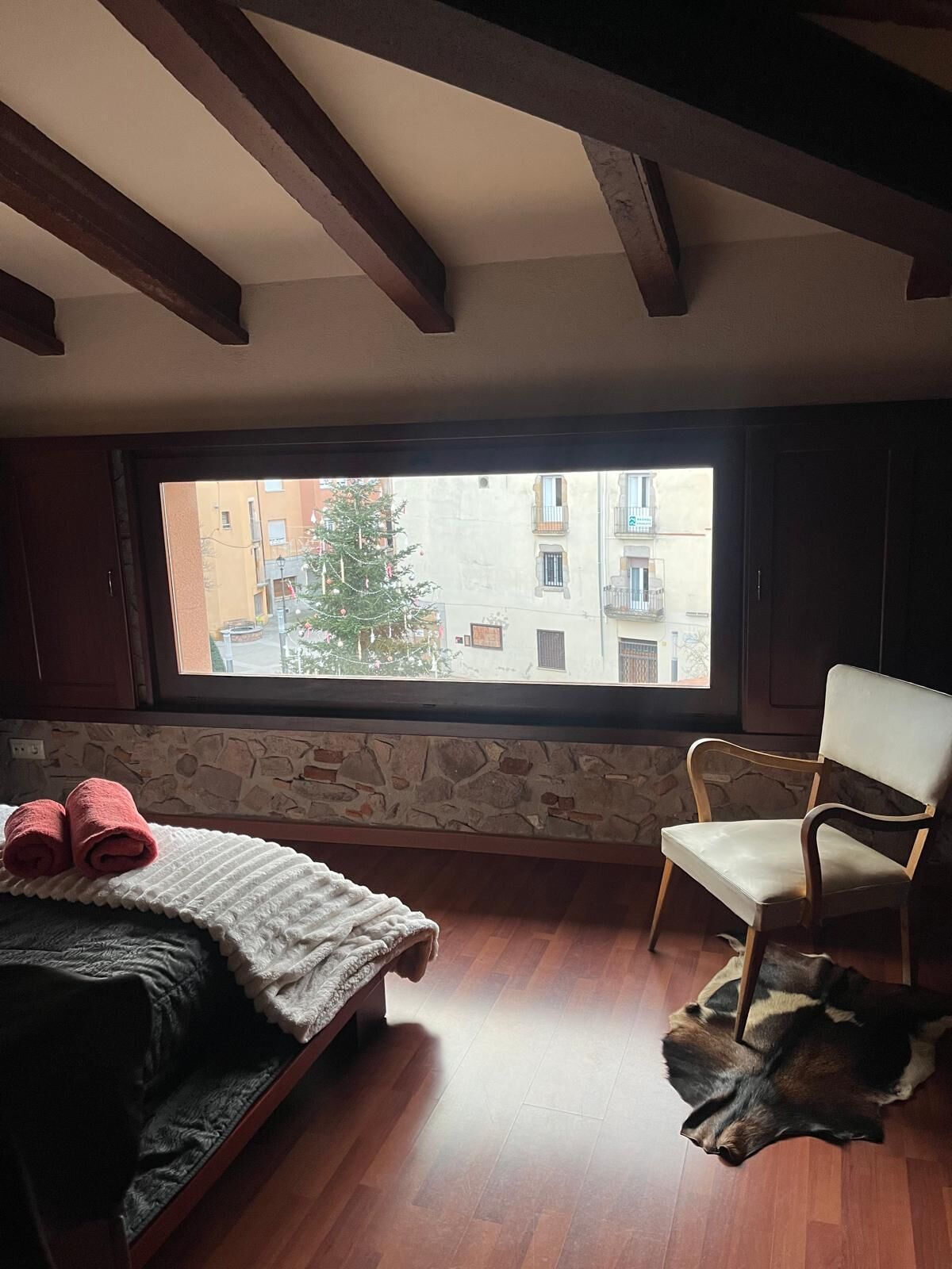 Une chambre avec un lit, une chaise, un mur en pierre et une fenêtre donnant sur des immeubles.