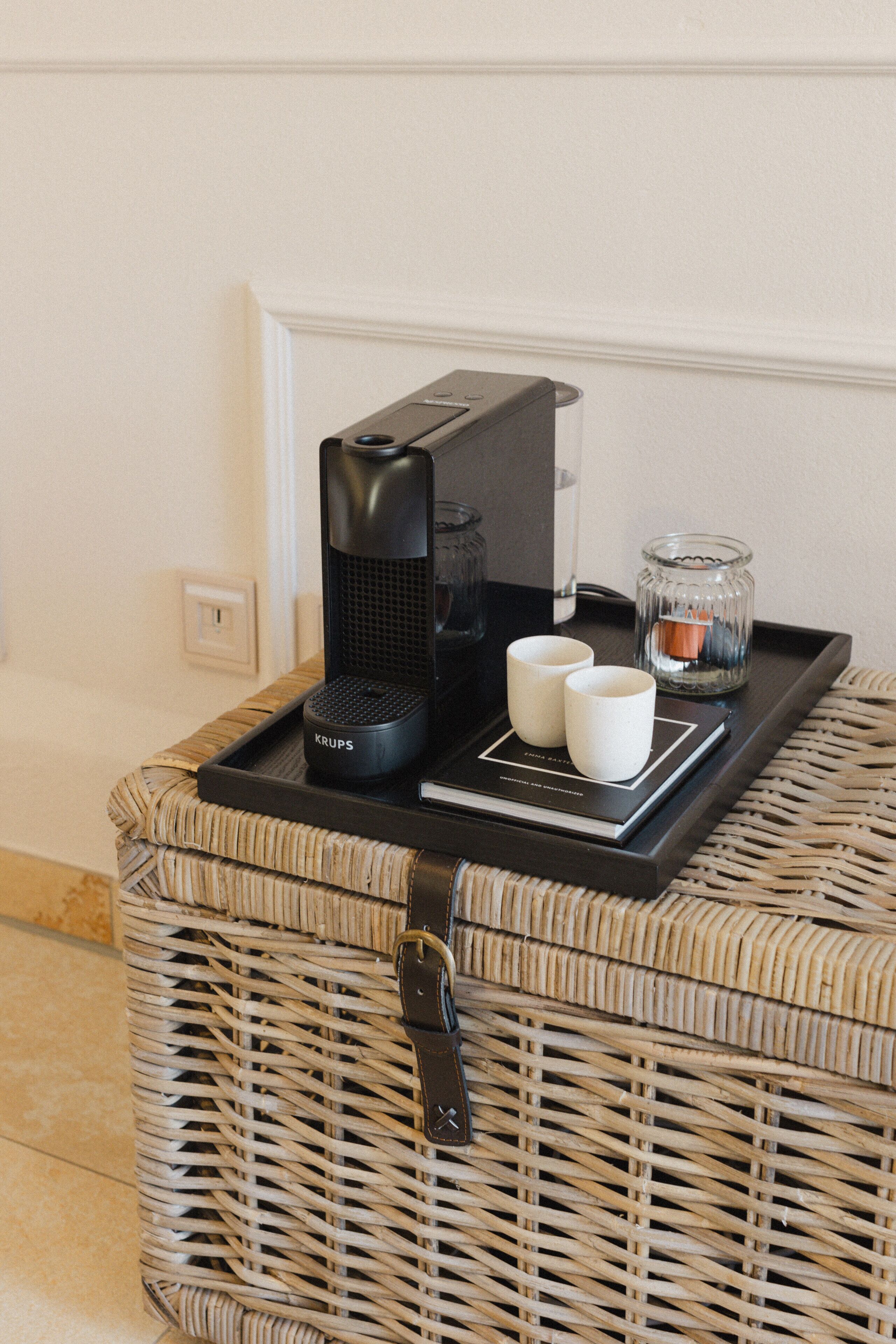 A wicker basket with a black coffee maker, two white cups, and a glass candle holder on a tray.