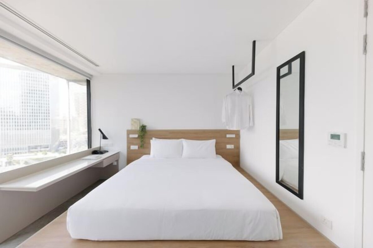 A modern hotel room with a large bed, a bedside table, a window with a view of the city, and a large mirror on the wall.