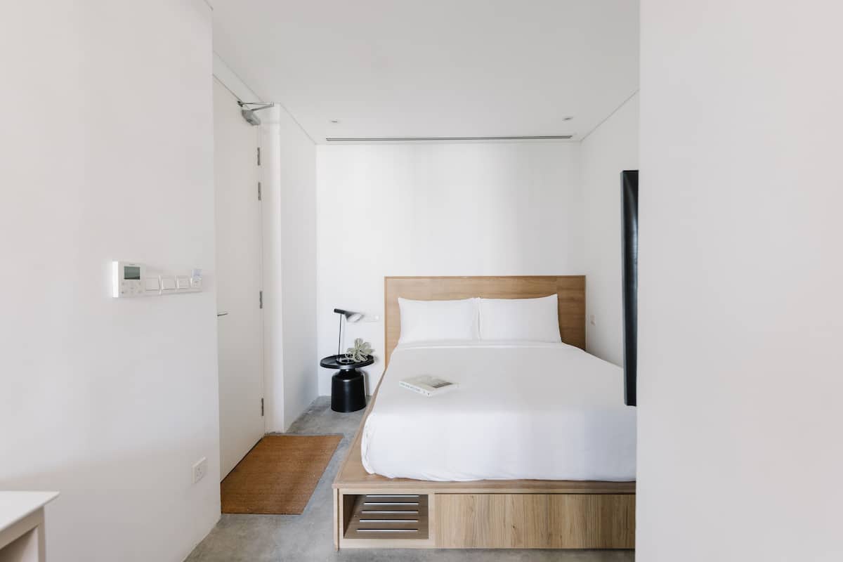 A modern bedroom with a wooden bed, white bedding, a bedside table, and a small black side table with a lamp.