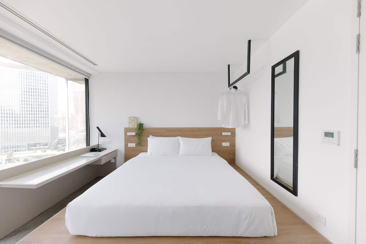 A modern hotel room with a large bed, a window with a view of the city, a bedside table with a lamp, and a mirror on the wall.