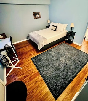 Ein Schlafzimmer mit einem Bett, einem Schreibtisch, einem Sessel, einem schwarzen Teppich mit Muster und einem kleinen gerahmten Bild an der Wand.