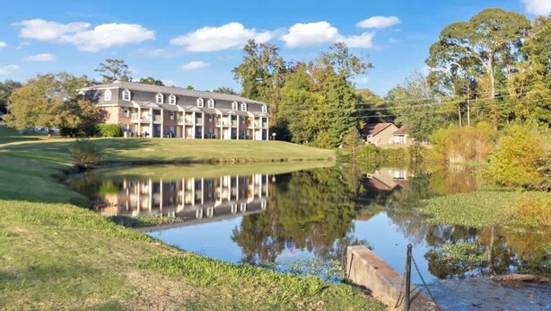 A multi-story building with a reflective pond in front, surrounded by greenery and trees. - AU Condo Getaway Across From Vet School - Kitchen, Pools, Queen BR, Pets Welcome (Auburn)