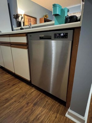 A modern kitchen with a stainless steel dishwasher, wooden cabinets, and a wooden floor. - AU Condo Getaway Across From Vet School - Kitchen, Pools, Queen BR, Pets Welcome (Auburn)