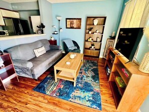 A living room with a grey sofa, a wooden coffee table, a blue patterned rug, a bookshelf, and a television. - AU Condo Getaway Across From Vet School - Kitchen, Pools, Queen BR, Pets Welcome (Auburn)