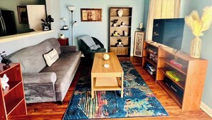 A living room with a grey sofa, a wooden coffee table, a bookshelf, a TV stand, and a colorful rug. - AU Condo Getaway Across From Vet School - Kitchen, Pools, Queen BR, Pets Welcome (Auburn)