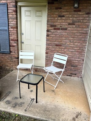 A patio with two white metal chairs and a black table, a white door, and a brick wall.