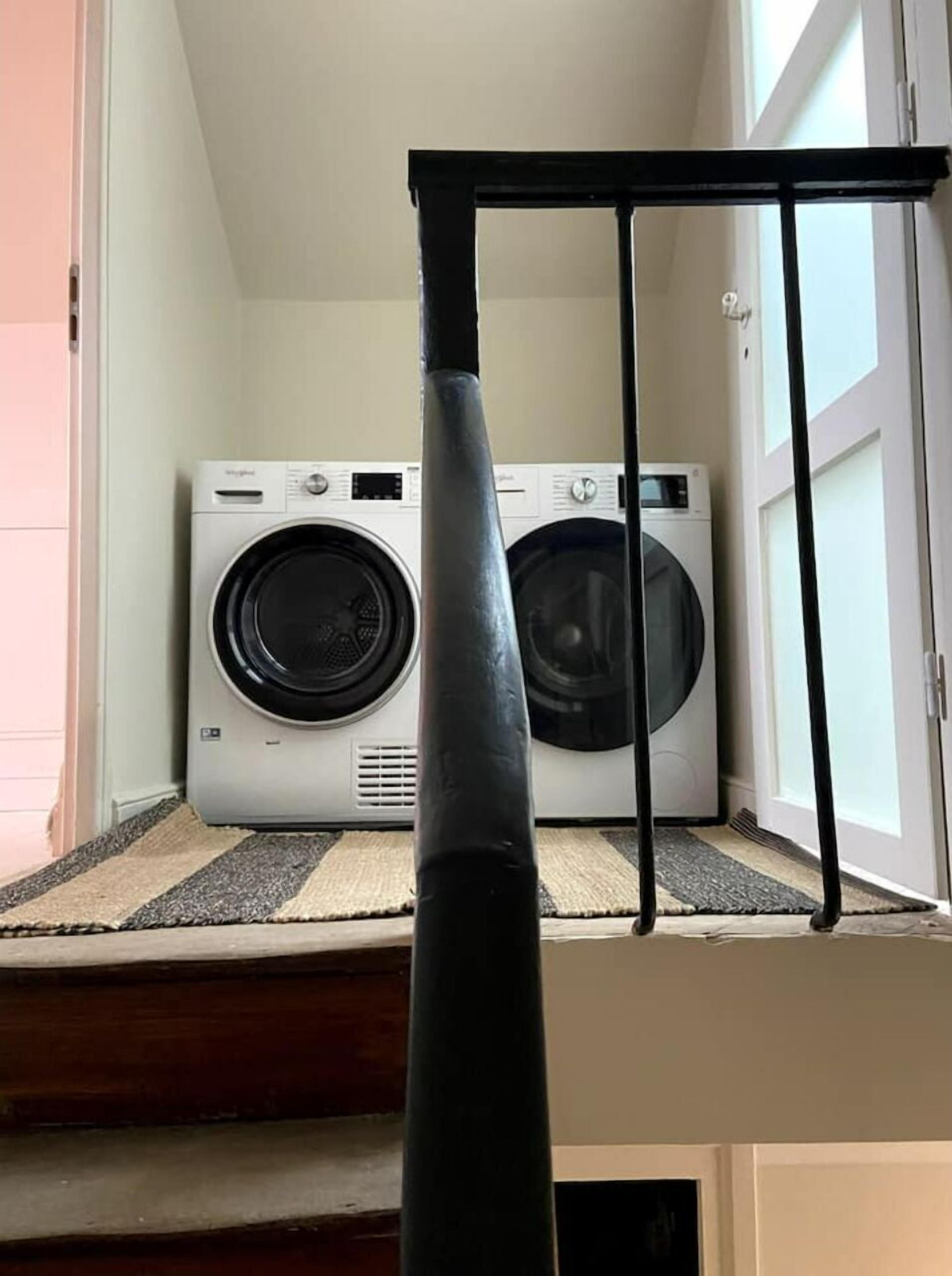 A laundry room with two front-loading washing machines and a metal drying rack in front of a window.