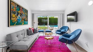 Een moderne woonkamer met een grijze bank, blauwe fauteuils, een kleurrijk vloerkleed en een groot schilderij.