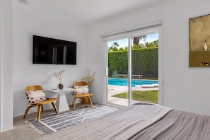 Quarto com cama grande, televisão na parede, mesa pequena com cadeiras e vista para uma piscina através de uma porta de correr de vidro.